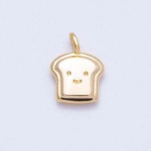 May include: A gold-colored pendant in the shape of a slice of bread with a smiling face. The pendant has a loop at the top for attaching to a chain or bracelet. The bread slice has a rounded top and a flat bottom.