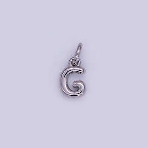 May include: A silver-colored letter "G" charm pendant. The charm is in a cursive style, with a rounded design. It has a small loop at the top for attaching to a necklace or bracelet. The background is a light purple color.