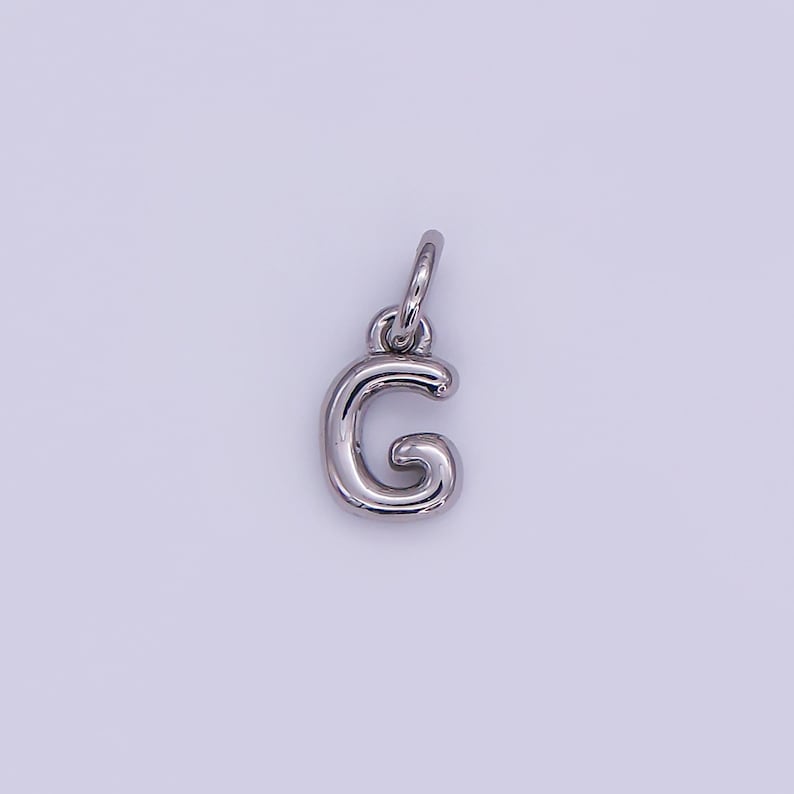 May include: A silver-colored letter "G" charm pendant. The charm is in a cursive style, with a rounded design. It has a small loop at the top for attaching to a necklace or bracelet. The background is a light purple color.
