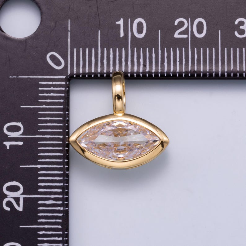 May include: A gold-toned pendant featuring a marquise-cut clear gemstone set in a smooth bezel. The pendant is positioned against a black ruler, showing a width of approximately 0.5 inches and a height of approximately 0.4 inches.