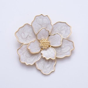 May include: A white and gold flower brooch with a marbled effect on the petals. The brooch has a gold-colored border and a cluster of small gold beads at the center, resembling a flower's stamen. The brooch is a jewelry item.
