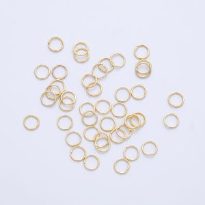 May include: A collection of small, gold-colored jump rings scattered on a white surface. The rings are circular and appear to be made of metal, likely for jewelry making or crafts. The lighting is bright, highlighting the metallic sheen.