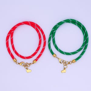 Puede incluir: Dos collares: uno rojo con detalles blancos, el otro verde con detalles blancos. Ambos tienen cierres dorados y dijes en forma de corazón. El diseño tipo cuerda añade una textura única a estos accesorios.