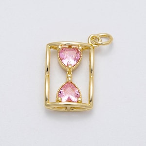 Puede incluir: Colgante dorado con forma de reloj de arena. El reloj de arena contiene dos gemas rosas en forma de corazón. El colgante tiene un marco rectangular y un lazo en la parte superior para una cadena.
