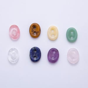 May include: Eight oval-shaped gemstone beads in various colors, including pink, brown, yellow, green, clear, blue, purple, and light pink. Each bead has two holes. The beads are arranged on a white surface.