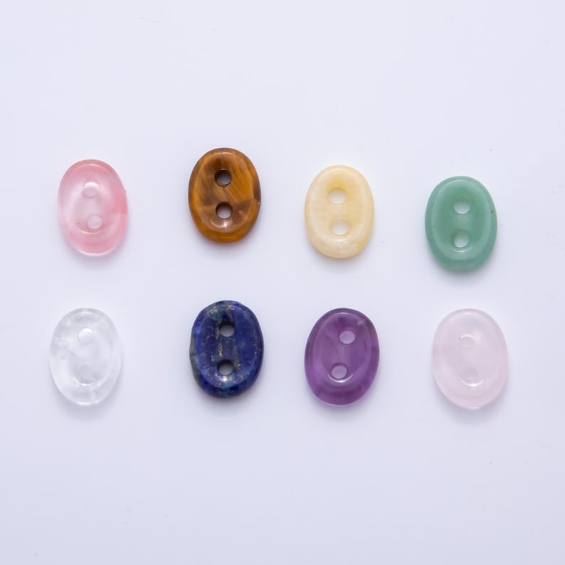 May include: Eight oval-shaped gemstone beads in various colors, including pink, brown, yellow, green, clear, blue, purple, and light pink. Each bead has two holes. The beads are arranged on a white surface.