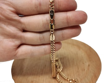 May include: A gold chain necklace with a curb link style. The chain is held in a hand, and the chain is laying on a wooden surface.