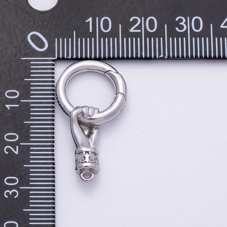 K&ouml;nnte beinhalten: Ein silberfarbener Anh&auml;nger mit einer Hand, die einen Ring h&auml;lt. Die Hand ist detailliert mit geformten Fingern und einer dekorativen Manschette. Der Ring ist an der Hand befestigt und hat einen Verschluss. Der Anh&auml;nger wird neben einem Lineal gezeigt.