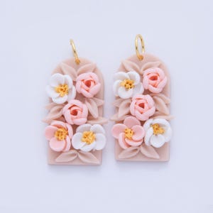May include: A pair of floral earrings. Each earring features a rectangular, blush-colored base adorned with white and pink flowers with yellow centers. Gold-colored hoops are attached at the top.