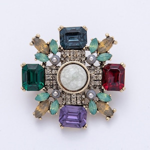 May include: A gold-toned brooch with a central white stone surrounded by a square pattern of clear crystals. The brooch features emerald green, deep red, purple, and teal-colored rectangular stones, along with smaller leaf-shaped accents.