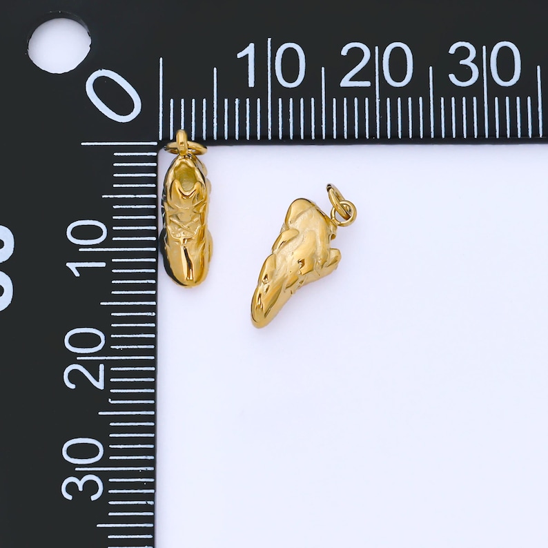 May include: Two gold-colored charms. One charm is shaped like a shoe, and the other is an abstract shape. Both charms have a small loop at the top for attaching to a chain or bracelet. The charms are displayed on a white surface next to a ruler.