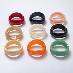 May include: Nine chunky resin rings in various colors, including red, navy, peach, tan, cream, black, yellow, orange, and green. The rings have a smooth, glossy finish and a thick band design, suitable for fashion accessories.