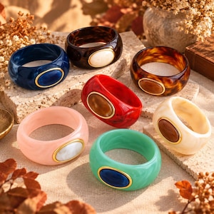 May include: A collection of six chunky resin bangle bracelets in various colors including navy, dark brown, red, light pink, mint green, and cream. Each bracelet features a smooth, rounded band with a central oval inlay framed in gold.