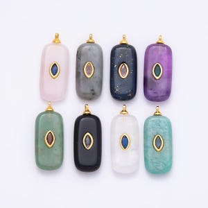 May include: Eight rectangular gemstone pendants in various colors, including pink, gray, navy, and purple. Each pendant has a gold-colored bail and a central oval-shaped gemstone in a contrasting color. The pendants are arranged on a white surface.