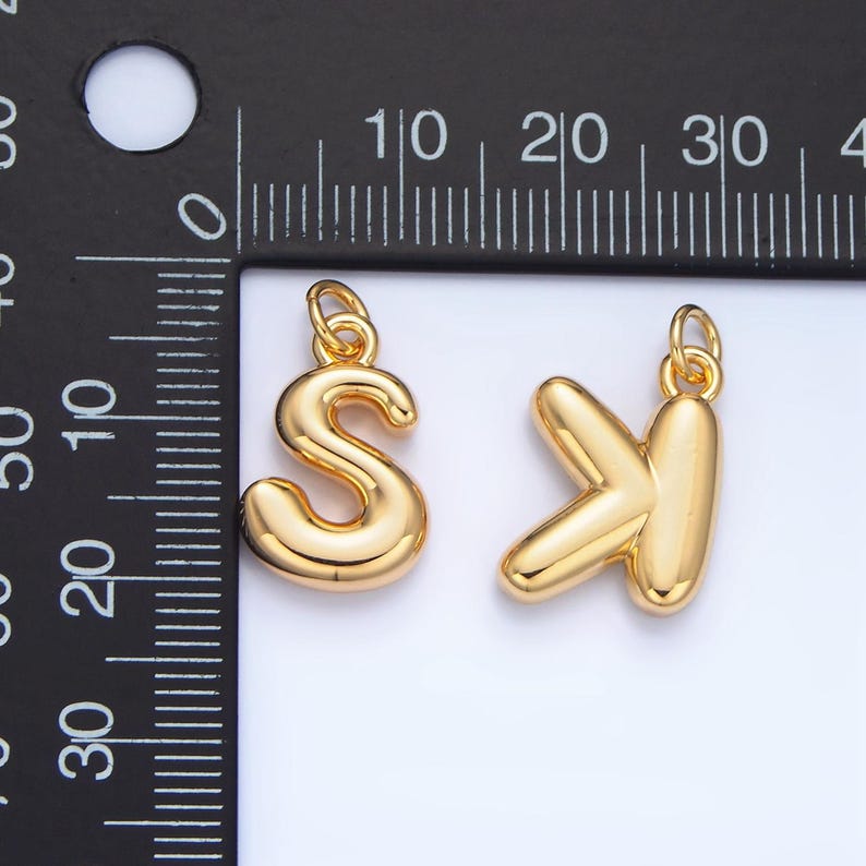 May include: Two gold-colored metal letter charms, one shaped like the letter "S" and the other shaped like the letter "Y". The charms are about 1 inch tall and are shown against a black ruler.