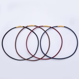 May include: Four circular cord necklaces arranged in a row, featuring black, brown, dark red, and navy blue colors. Each necklace is finished with a polished gold-tone metal cylindrical clasp mechanism that secures the ends of the cord together.