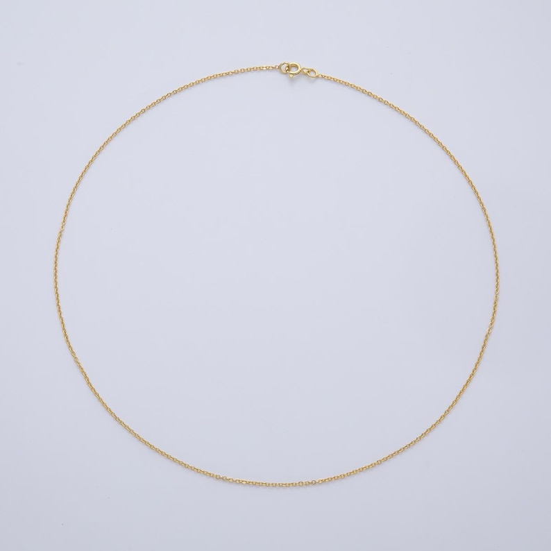 K&ouml;nnte beinhalten: Eine zarte, goldfarbene Halskette mit einem schlichten Kettendesign. Die Halskette wird vor einem schlichten, wei&szlig;en Hintergrund pr&auml;sentiert, der ihren minimalistischen Stil hervorhebt. Der Verschluss ist oben sichtbar.