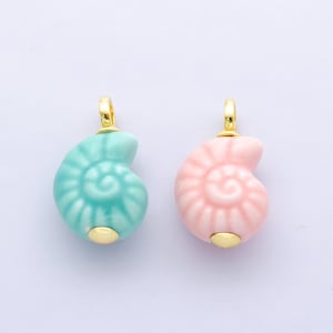 Puede incluir: Dos colgantes de cerámica con forma de concha. Uno es de color turquesa claro y el otro rosa. Ambos tienen una anilla dorada en la parte superior y una base dorada. Los colgantes son probablemente para hacer joyas.