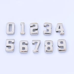 Può includere: Dieci numeri in metallo argentato, da zero a nove, disposti su uno sfondo bianco. Ogni numero ha un aspetto tridimensionale con una superficie lucida e riflettente. I numeri sono ben distanziati e chiaramente visibili.
