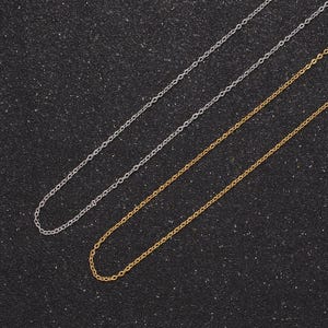 May include: Two chains, one silver and one gold, laid out on a black textured surface. The chains are both delicate and have a simple link design.