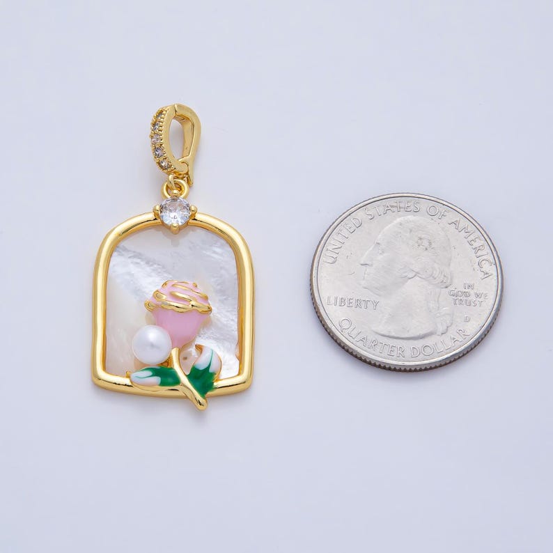 May include: Gold-tone pendant with a rectangular frame, featuring a pink rose, a white pearl, and green leaves. The pendant has a white background and a clear crystal at the top. A U.S. quarter is next to the pendant.