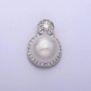 May include: A silver pendant featuring a large, lustrous white pearl encircled by a halo of small, sparkling clear stones. Above the pearl, a smaller, round cluster of similar stones surrounds a larger central stone.
