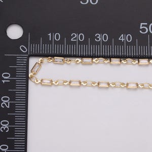 May include: Close-up of a gold-colored chain with rectangular, clear crystal accents. The chain is laid out on a white surface next to a black ruler with centimeter markings. The chain's design features small, rectangular crystals.