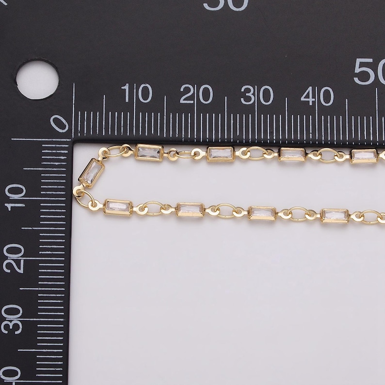 May include: Close-up of a gold-colored chain with rectangular, clear crystal accents. The chain is laid out on a white surface next to a black ruler with centimeter markings. The chain's design features small, rectangular crystals.