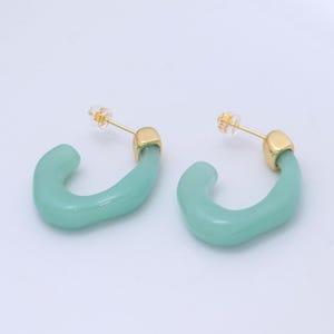 May include: A pair of mint green hoop earrings with gold-colored posts and accents. The earrings have a smooth, curved design, and are set against a light background.