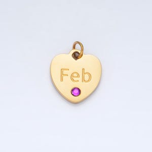 May include: Gold heart-shaped charm with the word "Feb" engraved on it. The charm has a small pink gemstone in the center.