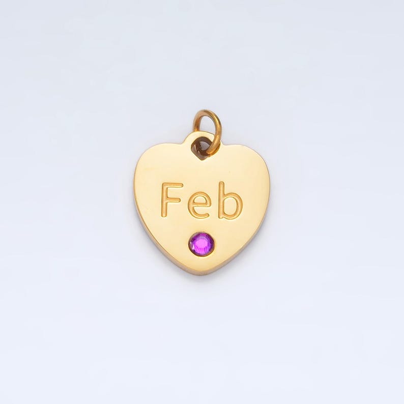 May include: Gold heart-shaped charm with the word "Feb" engraved on it. The charm has a small pink gemstone in the center.