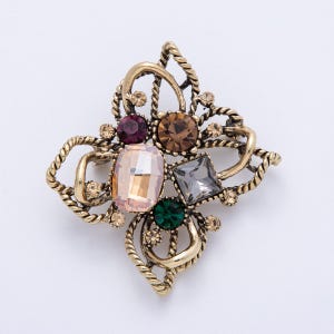 May include: A vintage-style brooch with a gold-tone metal base. The brooch features an ornate design with a central arrangement of colorful gemstones, including pink, brown, purple, green, and gray. Small, clear crystals are scattered around the larger stones.