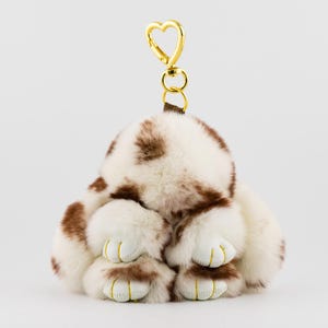 May include: A plush bunny keychain in white and brown spotted faux fur. The bunny has white paws with yellow stitching and a gold heart-shaped clasp. The keychain is designed to be a cute accessory.