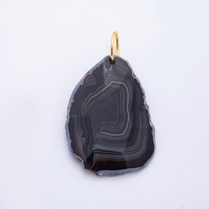 May include: A polished agate pendant with a gold-colored bail. The pendant is a dark gray color with lighter gray and white banding. The edges are a dark brown color with a blue-gray border. The pendant is irregularly shaped.