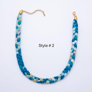 May include: Braided necklace in shades of blue, teal, and white with gold-tone chain and clasp. The necklace is labeled "Style # 2". The braided design adds texture and visual interest to this fashion accessory.