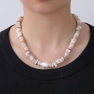May include: A necklace featuring a strand of small, irregular, off-white and tan shell pieces. The necklace has a large, white, baroque pearl pendant in the center. The necklace is worn against a black shirt.