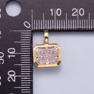 May include: A gold-toned rectangular pendant featuring a clear, faceted stone set in a smooth metal frame with a small bail at the top. The pendant is positioned against a black measuring tool with white markings, showing a width of 10 millimeters.