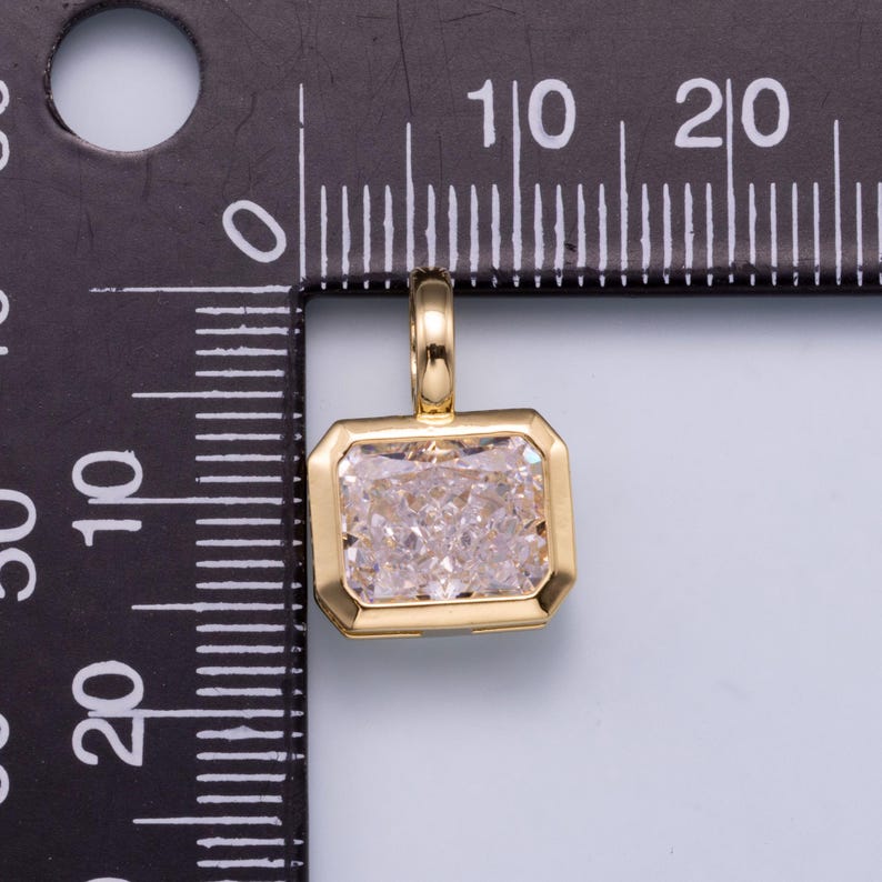 May include: A gold-toned rectangular pendant featuring a clear, faceted stone set in a smooth metal frame with a small bail at the top. The pendant is positioned against a black measuring tool with white markings, showing a width of 10 millimeters.