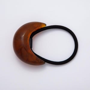 May include: A brown, crescent-shaped hair tie with a marbled effect. The hair accessory features a black elastic band and a decorative, curved accent. The hair tie is designed to hold hair in place and is suitable for various hairstyles.