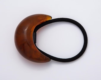 Op de afbeelding: Een bruine, halvemaanvormige haarband met een gemarmerd effect. Het haaraccessoire heeft een zwarte elastische band en een decoratief, gebogen accent. De haarband is ontworpen om het haar op zijn plaats te houden en is geschikt voor verschillende kapsels.