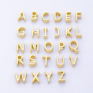 May include: Gold-colored alphabet letter charms, capital letters A through Z, arranged on a white surface. Each letter is a separate charm, suitable for jewelry making or craft projects. The letters are in a sans-serif font.