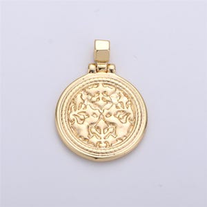 May include: A gold-toned pendant with a circular design featuring an intricate floral pattern. The pendant has a loop at the top for attaching to a chain.