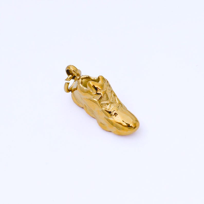 May include: A gold-colored sneaker pendant with intricate detailing. The pendant is a miniature replica of a running shoe, featuring laces and a textured sole. The overall design is shiny and reflective, suggesting a polished finish.
