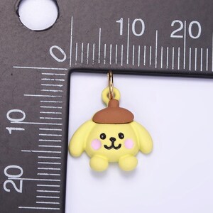 May include: A yellow cartoon dog charm with a brown hat, pink cheeks, and black eyes. The charm is attached to a gold jump ring.