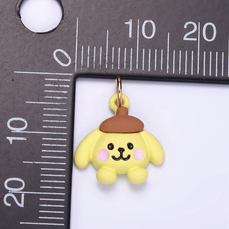 May include: A yellow cartoon dog charm with a brown hat, pink cheeks, and black eyes. The charm is attached to a gold jump ring.