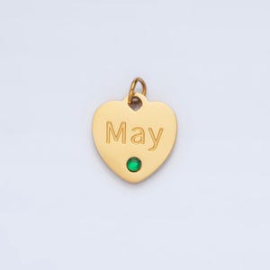 May include: Gold heart-shaped pendant with the word "May" engraved on it. A small green gemstone is set in the center of the pendant.