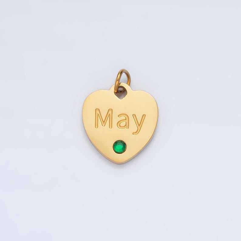 May include: Gold heart-shaped pendant with the word "May" engraved on it. A small green gemstone is set in the center of the pendant.