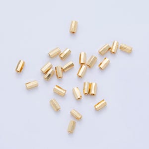 May include: A collection of small, gold-colored, hexagonal beads scattered on a white surface. Each bead has a cylindrical shape with multiple flat sides, reflecting light. The beads appear to be made of metal.
