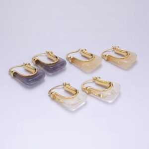 May include: Six pairs of gold-tone hoop earrings with square, translucent, gemstone-like pendants. The pendants are in shades of purple, yellow, and white. The earrings are displayed on a white surface.