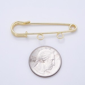May include: A gold-toned metal safety pin featuring two small circular loops attached to the bottom bar. The safety pin is positioned above a standard United States quarter coin, which serves as a visual reference for its size and scale.